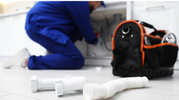 Plumber inspecting plumbing under a kitchen sink while disconnected PVC pipes rest on the floor during a potential broken pipe emergency